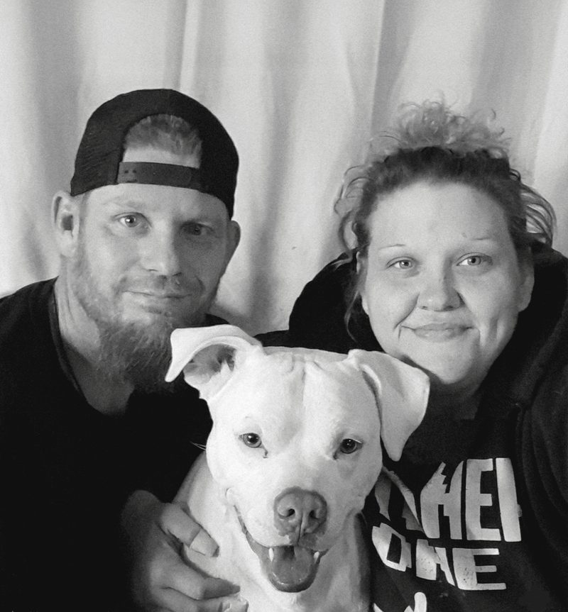 Black-and-white photo of Nathan in a backwards cap with Stephanie, both smiling, and their white dog Rebel between them